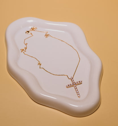 Gold necklace with a cross pendant on a white dish against a beige background