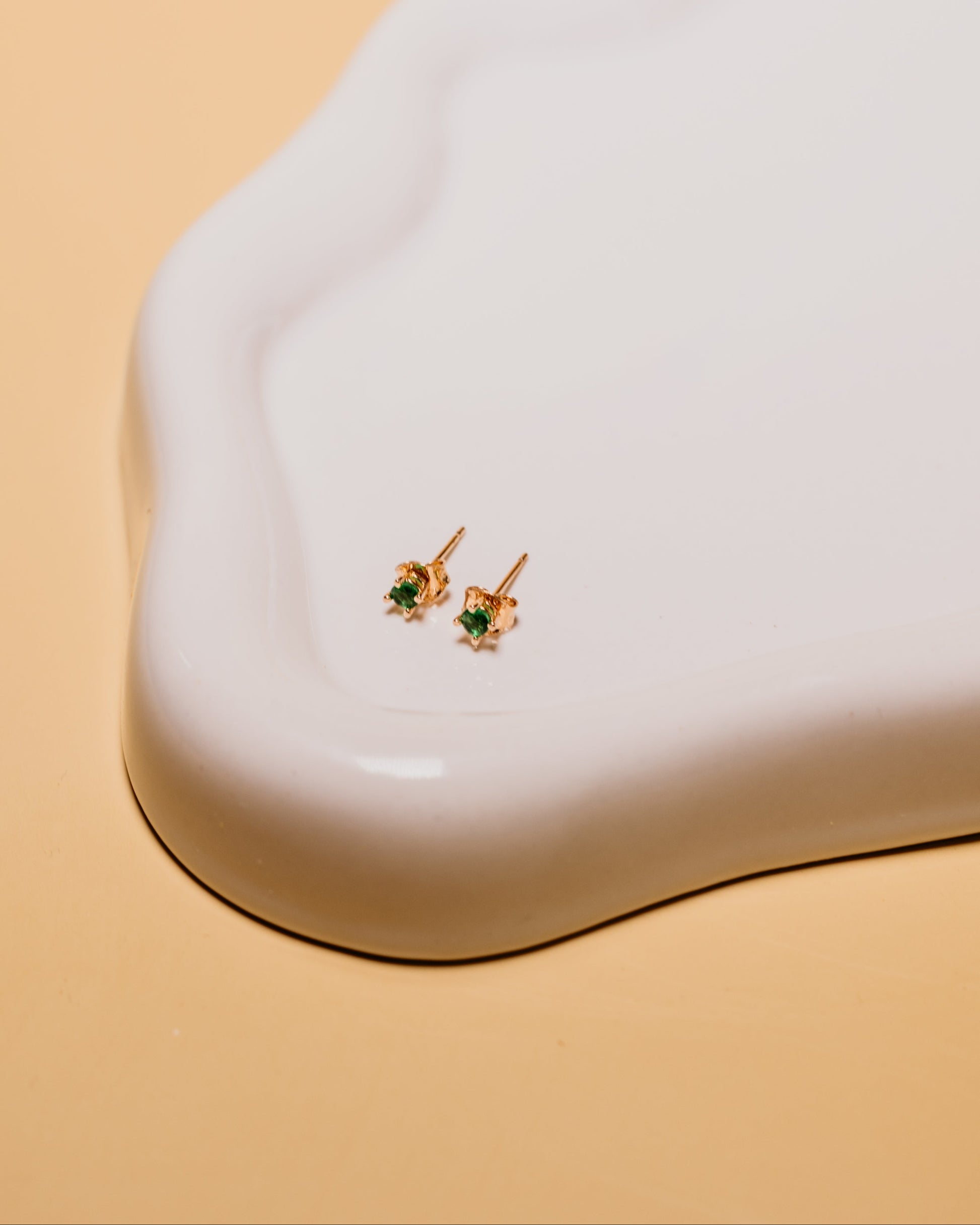 Gold earrings with green gemstones on a white stand against a beige background