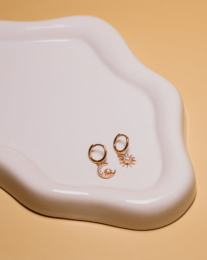Rose gold earrings with moon and star designs on a white dish against a beige background