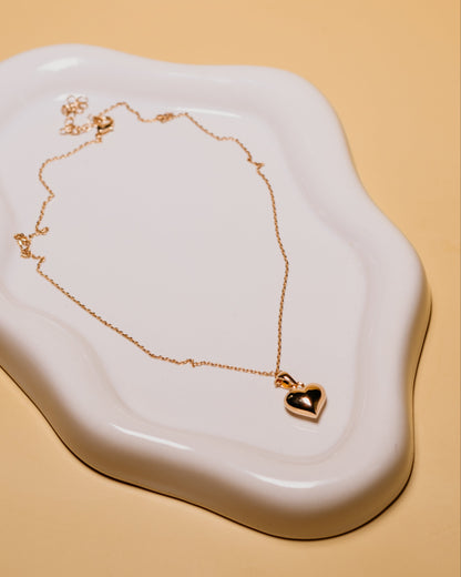 Gold necklace with a heart pendant on a white dish against a beige background